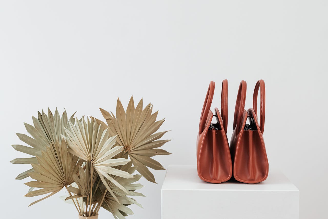 discover Elegant brown handbags alongside dried palm leaves on white background.